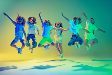 Group of children, little girls in sportive casual style clothes dancing in choreography class isolated on green background in yellow neon light. Concept of music, fashion, art