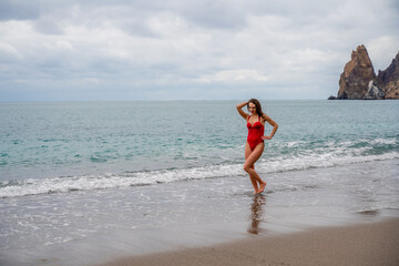 A beautiful and sexy brunette in a red swimsuit on a pebble beach, Running along the shore in the foam of the waves