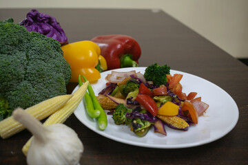 Picture of sautéed salad on a white plate along with healthy raw boiled exotic vegetables. baby corn, capsicum, bell pepper, broccoli, garlic, chilly, cabbage, tomato, onion, carrot on wooden table.