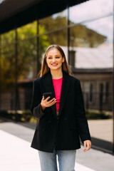 Fototapeta premium young business woman goes and uses mobile phone in her hands a urban background a modern office building.