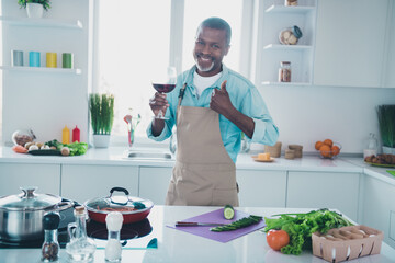 Photo of confident pretty mature guy dressed apron showing thumb up drinking wine indoors home room