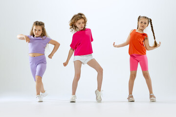 Fototapeta premium Three little girls, kids in bright colorful clothes dancing, posing isolated on white studio background. Concept of music, fashion, art, childhood, hobby