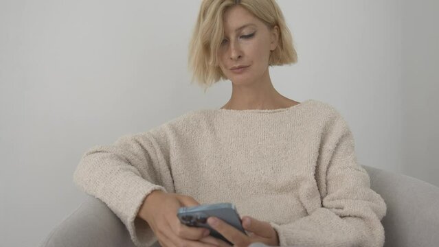 Woman holding in hands and using smartphone in the living white room at home, typing message browsing internet. Sitting on armchair 