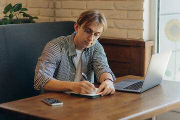 Smiling man with glasses sitting at table in cafe with laptop computer and writing something
