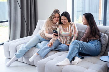 Obraz premium Happy young carefree diversity women friends in casual using phone, having fun together at home