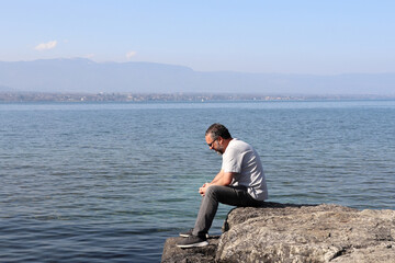 Naklejka na ściany i meble A charming man about 40 years old, sitting on rocks staring out to the lake.