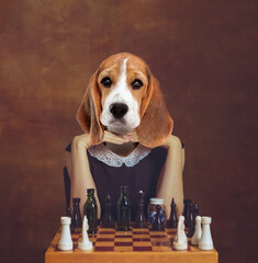 Young woman with dog's head sitting and playing chess isolated on brown background. Retro style, comparison of eras concept. Surrealism