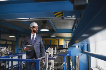 Chief Engineer in the hard hat walks through industrial factory while holding tablet.