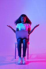 Shocked African American woman using laptop pc, sitting on chair, opening mouth in surprise in neon light, full length
