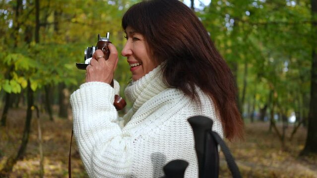 Side view curios talented mature woman taking photos with camera standing in autumn forest outdoors. Smiling happy Caucasian retiree enjoying hobby outdoors in park with poles for nordic walking aside