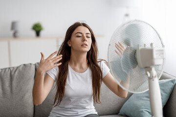Sad young european woman suffers from unbearably too hot weather, catches cold air from fan in living room - obrazy, fototapety, plakaty