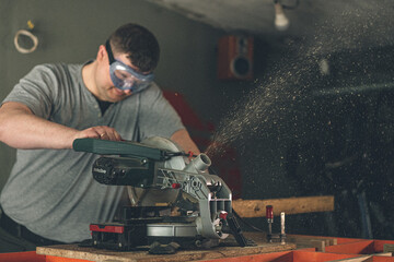 Strong male carpenter at work. The joiner holds the miter saw and a piece of plywood with his hand. Eyes are protected by special glasses. - obrazy, fototapety, plakaty