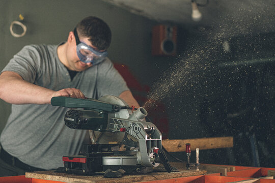 Strong male carpenter at work. The joiner holds the miter saw and a piece of plywood with his hand. Eyes are protected by special glasses.