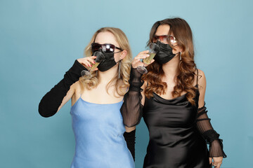 2 Mädchen in eleganten Kleidern und mit Sonnenbrillen, trinken Champagner durch schwarze medizinische Masken hindurch.  Studio Close-up, freigestellt  vor hellem Hintergrund