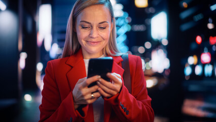 Portrait of a Beautiful Woman in Red Coat Walking in a Modern City Street with Neon Lights at Night. Attractive Female Using Smartphone and Looking Around the Urban Cinematic Environment.