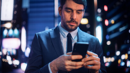 Portrait of a Handsome Man in Stylish Suit Walking in a Modern City Street with Neon Lights at Night. Attractive Male Using Smartphone and Looking Around the Urban Cinematic Environment.