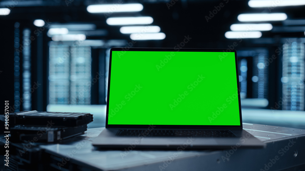 Poster Laptop Computer with Green Screen Chroma Key Mock Up Display Stands on a Table. In the Background Data Center With Rows of Server Racks and IT Engineer Working. Advanced Equipment.