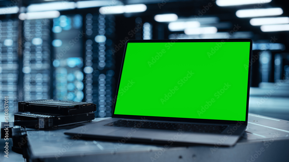 Poster Laptop Computer with Green Screen Chroma Key Mock Up Display Stands on a Table. In the Background Data Center With Rows of Server Racks and IT Engineer Working. Advanced Equipment.