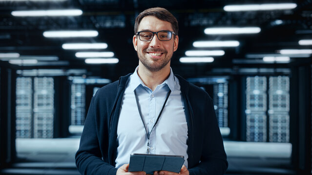 Portrait of a Bearded Handsome Caucasian IT Specialist in Glasses Standing with Tablet and Posing in the Middle of a Working Data Center Server Room with Server Computers Working on a Rack.