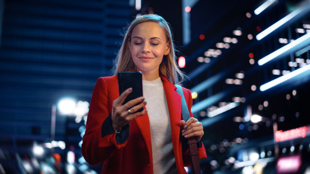 Portrait of a Beautiful Woman in Red Coat Walking in a Modern City Street with Neon Lights at Night. Attractive Female Using Smartphone and Looking Around the Urban Cinematic Environment.