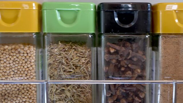 Closeup dolly shot of a range of cooking herbs and spices in glass storage jars with plastic lids, side-by-side in a wall rack.