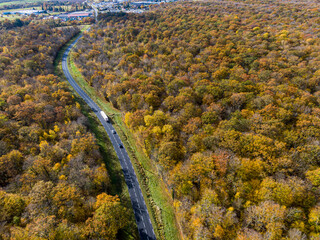 Forest road with white truck during Autumn. Aerial view road crossing a yellow and gold deciduous trees forest, Autumn