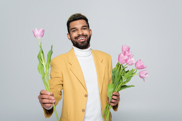 joyful african american man in stylish outfit holding bouquet of pink tulips isolated on grey.