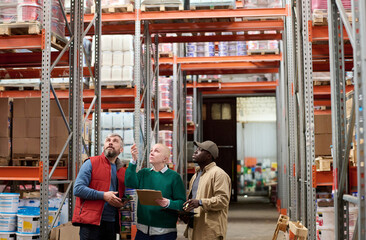 Woman pointing up on the shelves and counting goods together with her colleagues while they standing in storage room