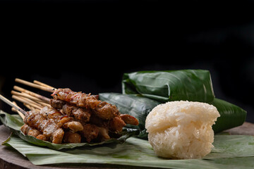 front view of grilled skewered pork(Moo Ping) with white sticky rice and sticky rice wrapped in banana leaves placed on the banana leaves and wood floor, black background