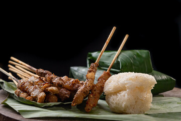 front view of grilled skewered pork(Moo Ping) with white sticky rice and sticky rice wrapped in banana leaves placed on the banana leaves and wood floor, black background