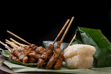 front view of grilled skewered pork(Moo Ping) with white sticky rice and sticky rice wrapped in banana leaves placed on the banana leaves, black background