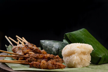 front view of grilled skewered pork(Moo Ping) with white sticky rice and sticky rice wrapped in banana leaves placed on the banana leaves and wood floor, black background