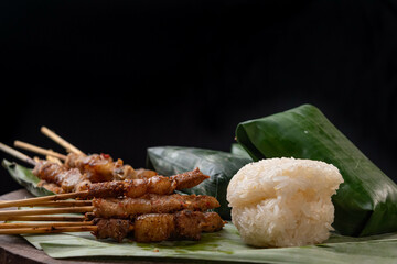 front view of grilled skewered pork(Moo Ping) with white sticky rice and sticky rice wrapped in banana leaves placed on the banana leaves and wood floor, black background