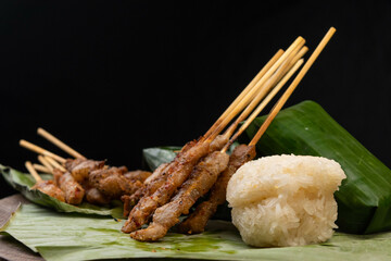 front view of grilled skewered pork(Moo Ping) with white sticky rice and sticky rice wrapped in banana leaves placed on the banana leaves and wood floor, black background