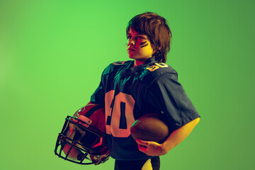 Closeup sportive strong kid, american football player in blue sports uniform and protective equipment posing isolated on green background in neon light.