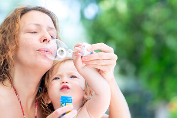 Obraz premium Little girl plays with soap bubbles. Mom and daughter by the pool. Fun summer day with family. Happy child blowing bubbles