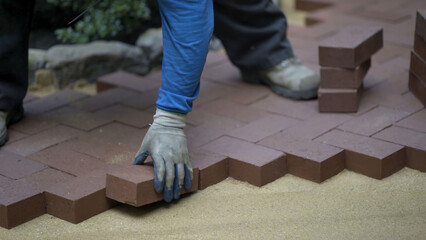 Hand putting red brick pavers into place in a herringbone pattern on a bed of sand in hardscaping landscaping patio project.