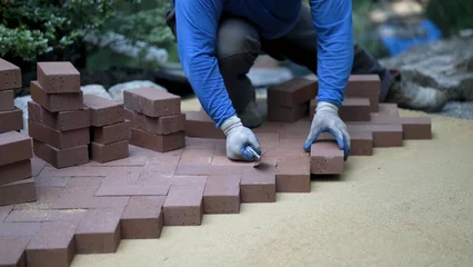 Tuinposter Using the butt of his trowel, a hardscaper taps the red brick pavers into place on a bed of sand in hardscaping landscaping patio project. © Robert Peak