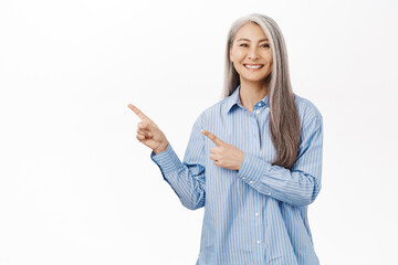 Smiling asian woman looking pleased, showing advertisement, pointing left, standing against white...