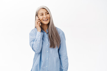 Portrait of happy smiling asian senior woman, old lady using smartphone, talking on mobile phone, standing with cellphone against white background