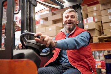 Portrait of warehouse worker smiling at camera while operating a forklift, he load cardboard boxes...