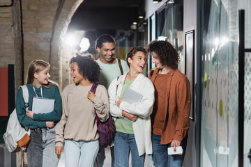 Fototapeta premium cheerful interracial students with backpacks smiling at each other in hallway of university.