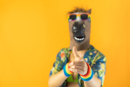 Portrait of lesbian female wearing crazy horse animal mask and LGBTQ+ pride rainbow wristband and sunglasses pointing at the camera isolated on orange background