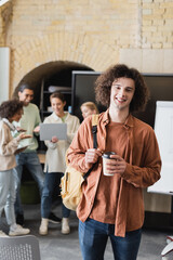 curly student with backpack and takeaway drink near blurred friends looking at laptop.