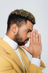 side view of young african american man in stylish yellow blazer with praying hands isolated on grey.