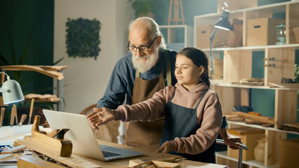Grandfather Carpenter with his Teenage Granddaughter Develop Wood Layouts, Model 3D Sketches Using a Laptop in the Workshop. Communication of Generations. Craft, Joinery and New Computer Technologies.