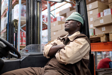 African young driver of forklift sitting on seat and relaxing after work at warehouse