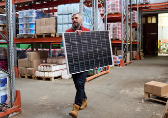 Mature bearded loader walking along the shelves with parcels with a solar battery in his hands in warehouse