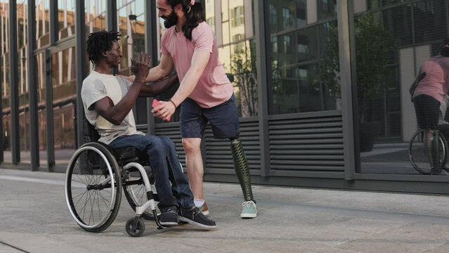 Multiracial people with disability hugging each other outdoor - Man with leg prosthesis greeting friend sitting on wheelchair
