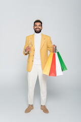 full length of happy african american man in yellow blazer holding shopping bags and glass of champagne on grey.